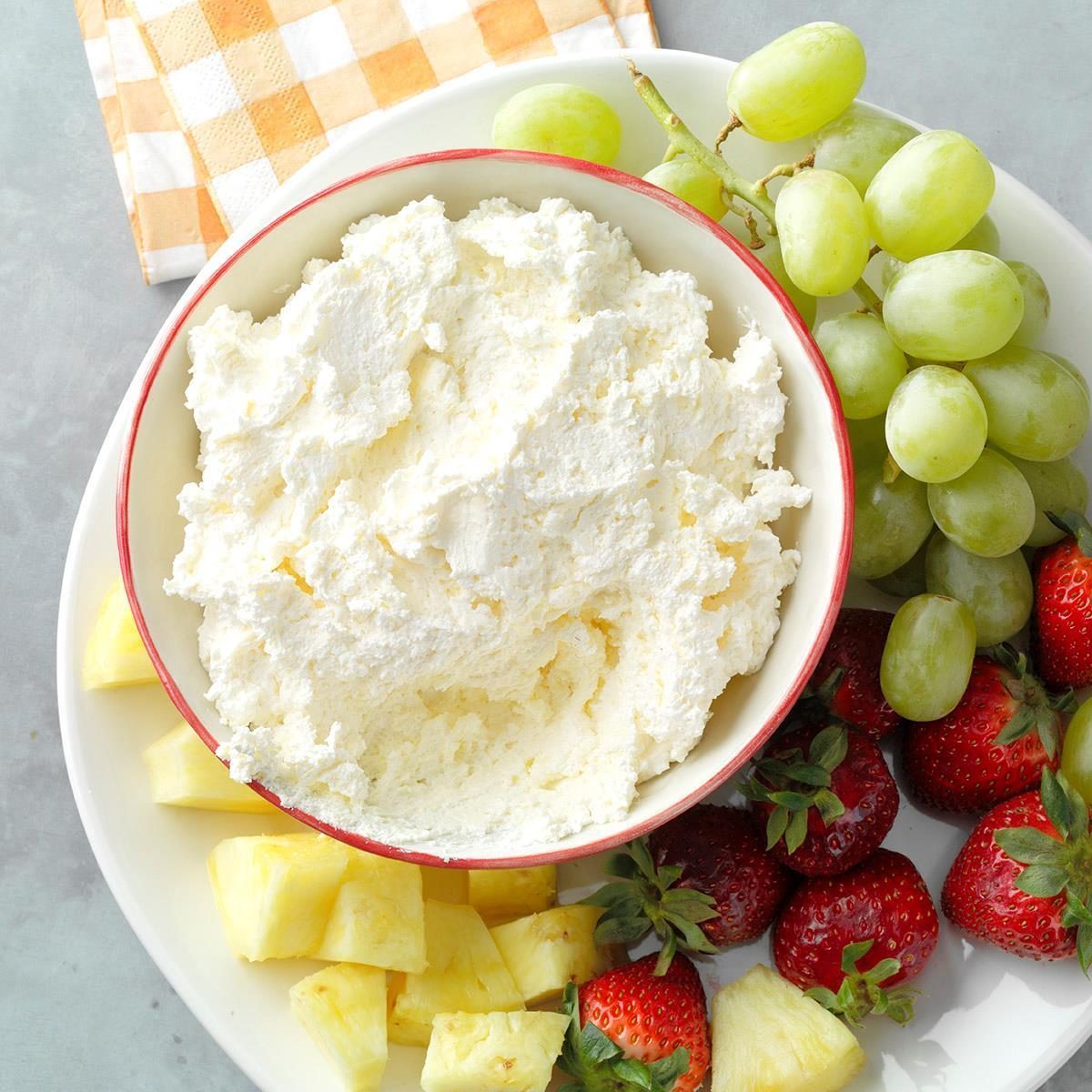 Bowl of whipped cream sits surrounded by green grapes, strawberries, pineapple chunks on a white plate, with a checkered napkin nearby.