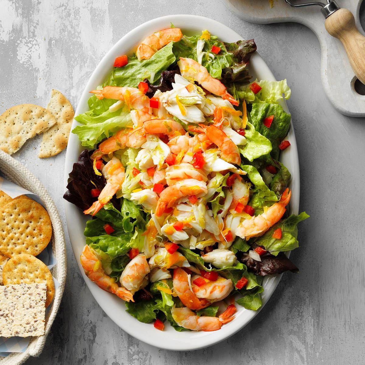 Salad with shrimp and crab sits on a white plate, garnished with red peppers, surrounded by crackers on a light gray surface.