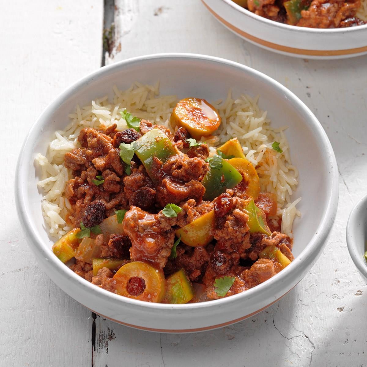 Bowl of rice topped with seasoned minced meat, sliced green olives, bell peppers, garnished with cilantro, placed on a white wooden table.