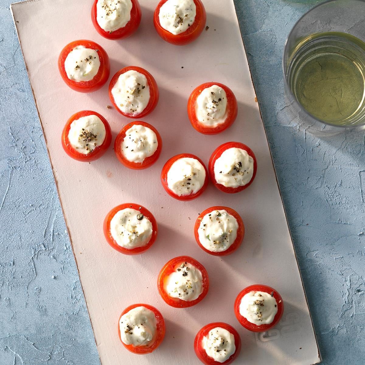 Stuffed cherry tomatoes sit on a cutting board, filled with creamy topping, sprinkled with black pepper; a glass of white wine nearby.