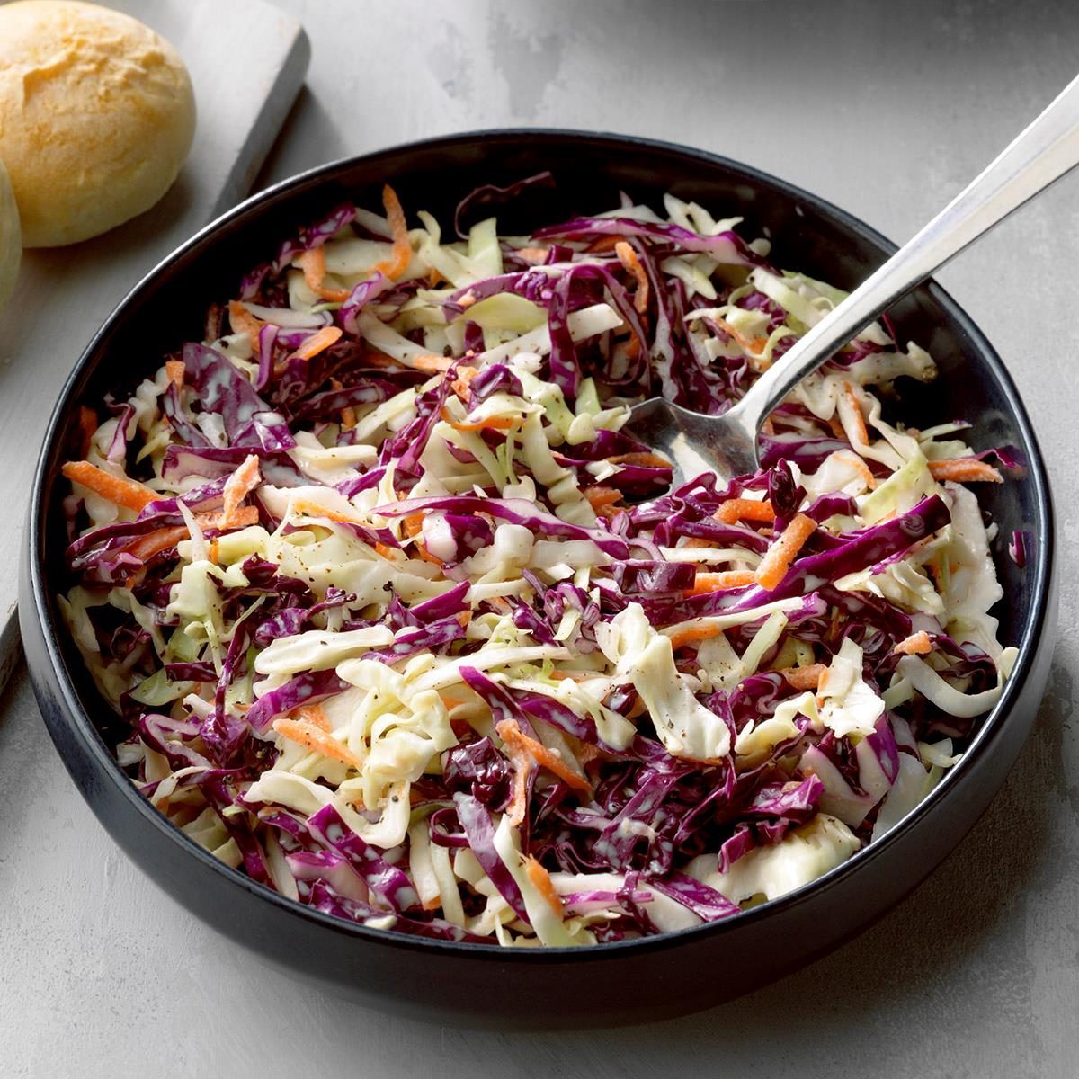 Coleslaw, mixed with dressing, sits in a black bowl. Nearby lies a roll on a gray surface.