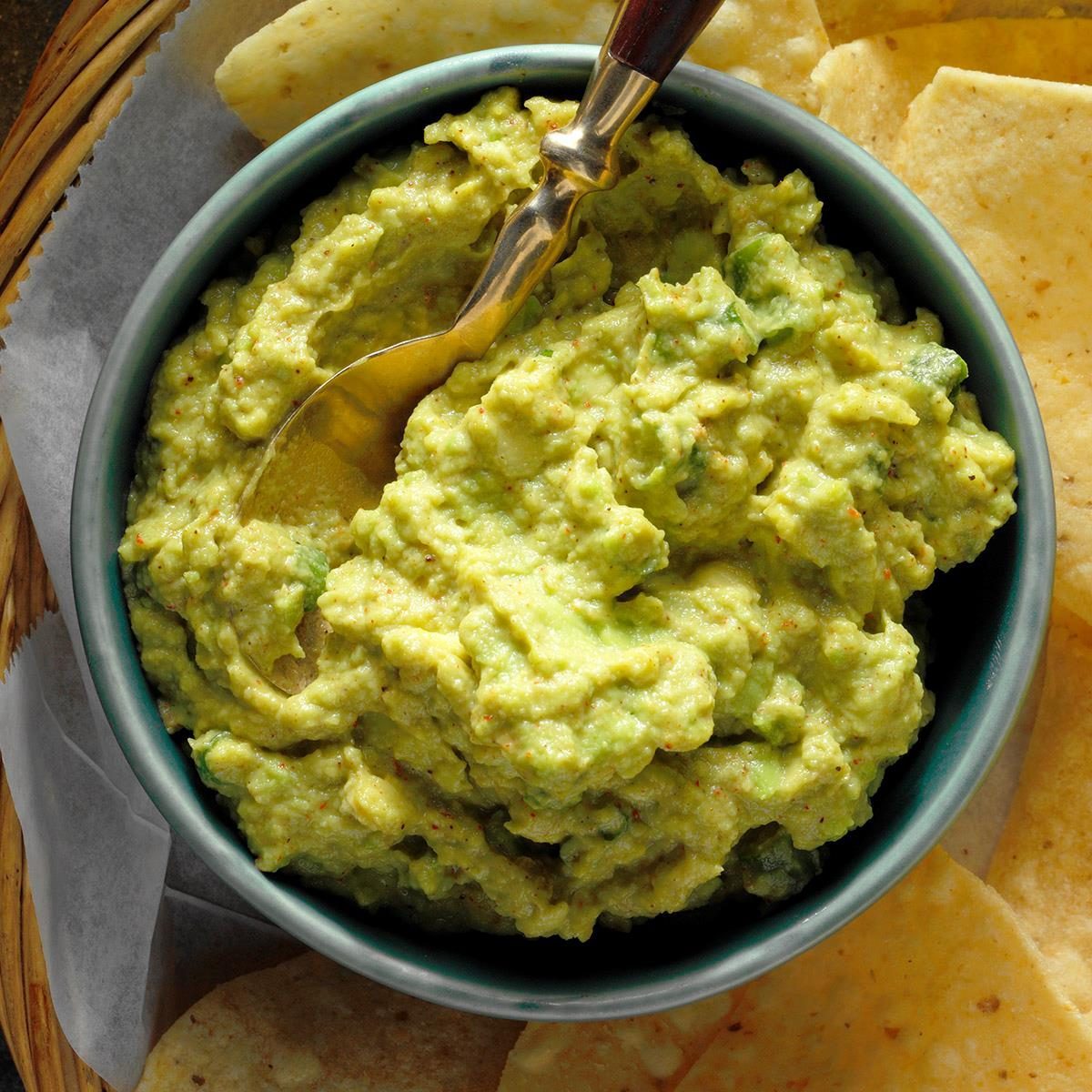 A bowl of guacamole sits beside tortilla chips on a wooden tray, with a spoon resting inside the dip.