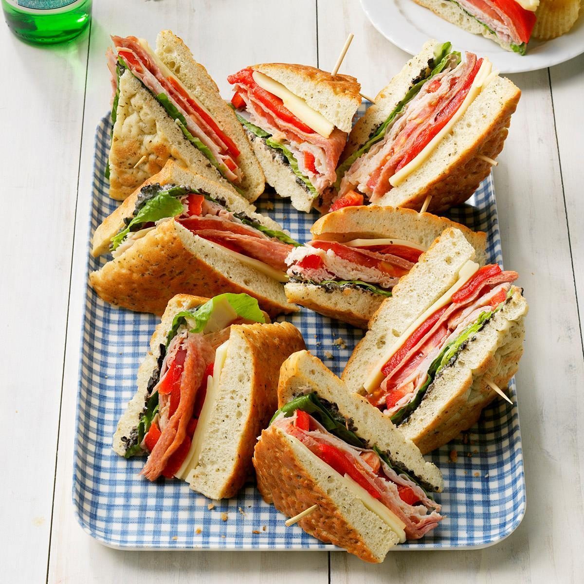 Sandwiches are arranged on a blue checked platter, filled with meats, cheese, and lettuce, set on a white wooden surface.