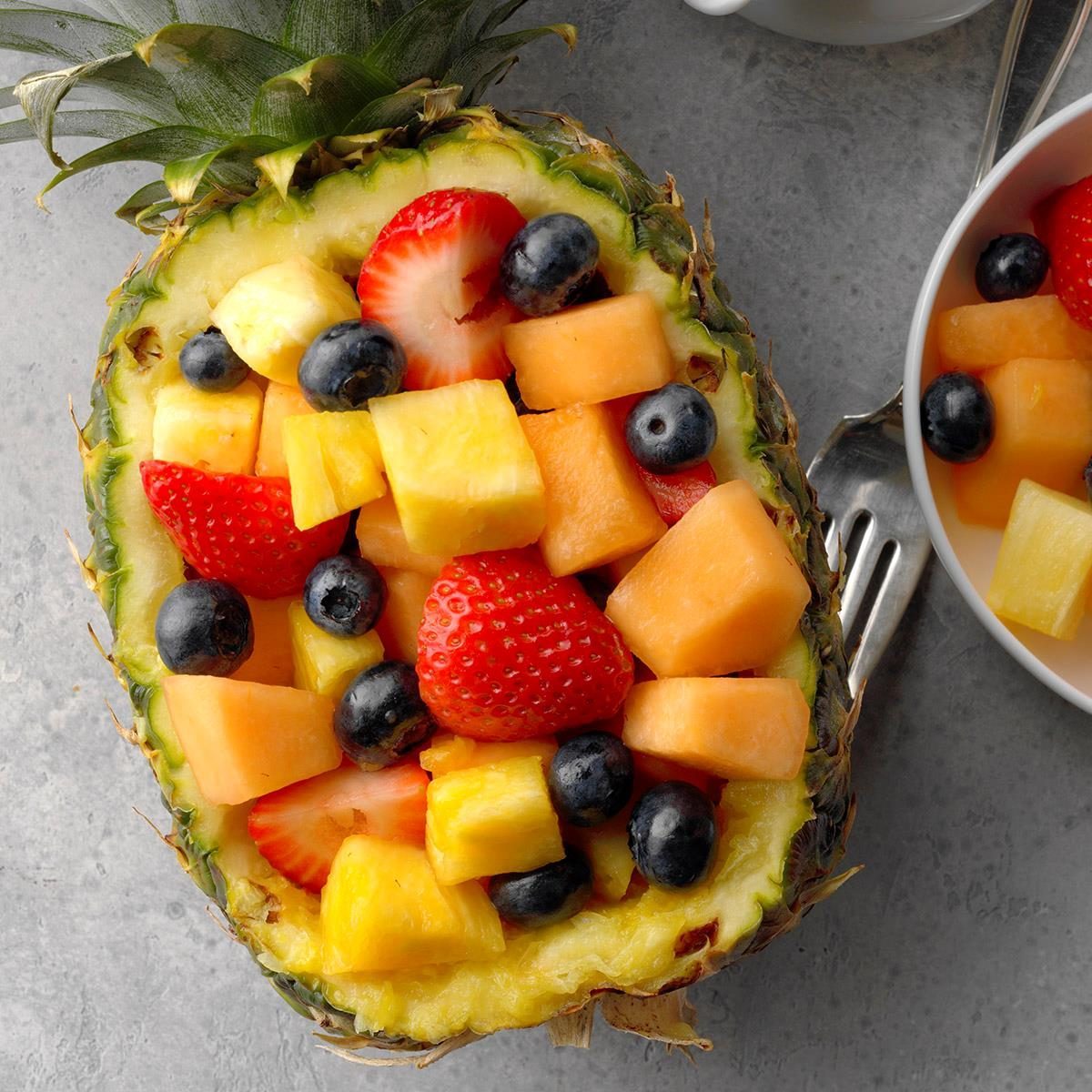 Pineapple bowl filled with colorful fruit pieces, surrounded by a fork and a partial view of a bowl on a gray surface.