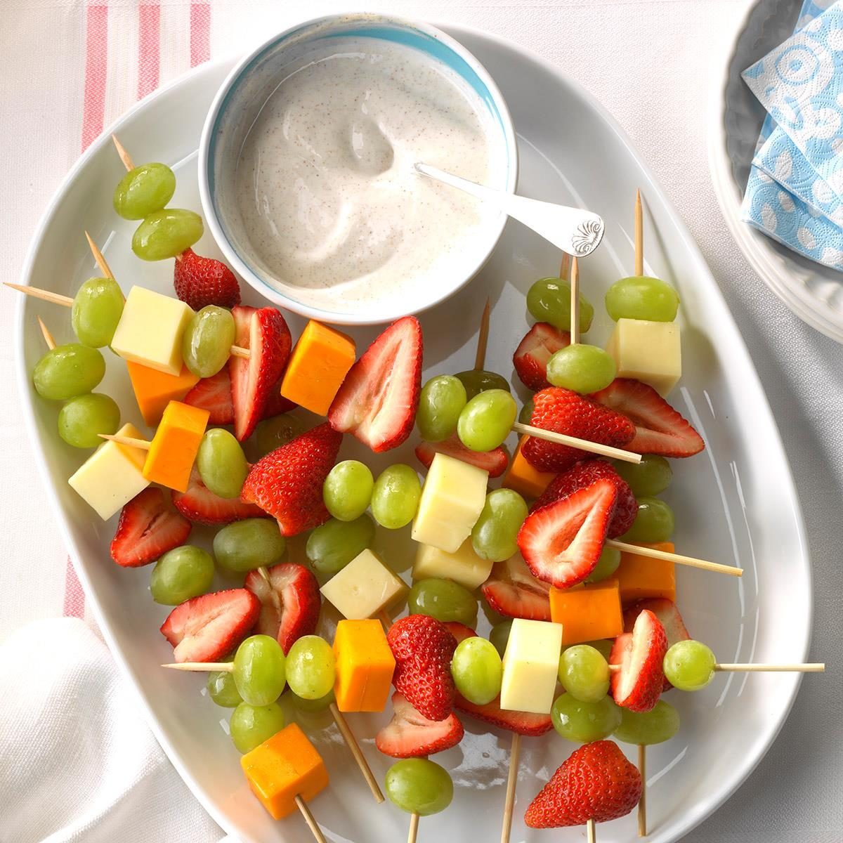 Skewers with grapes, strawberries, and cheese rest on a platter beside a bowl of creamy dip; napkins are nearby.