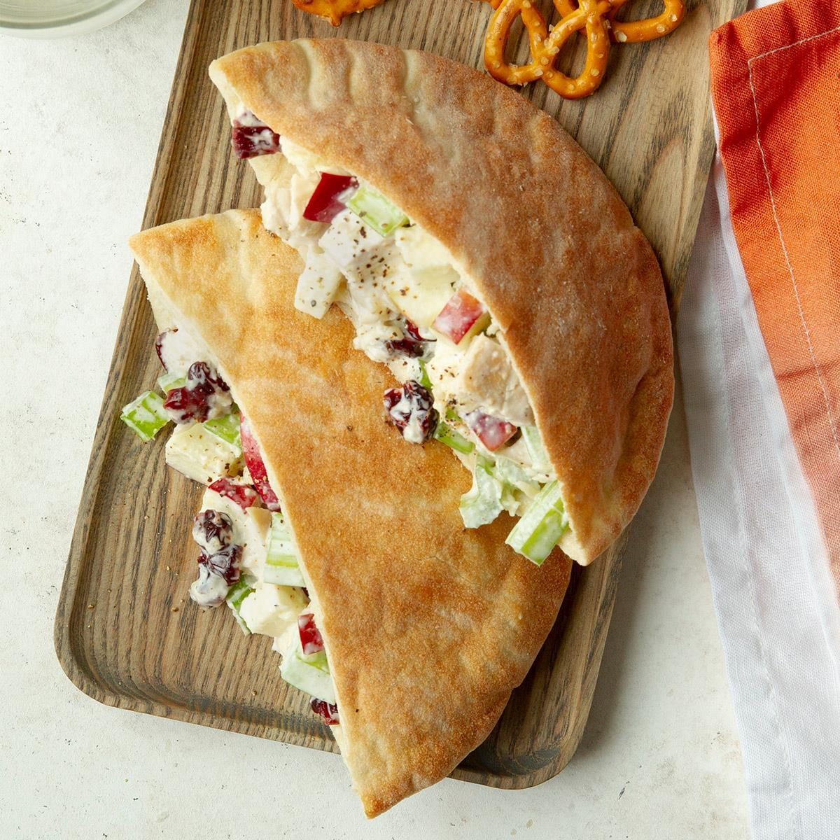 Pita pocket filled with chicken salad, apples, and celery is placed on a wooden tray, next to pretzels and an orange napkin.
