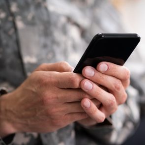 Hands hold a black smartphone, texting or browsing, dressed in a camouflage-patterned uniform, suggesting a military context.