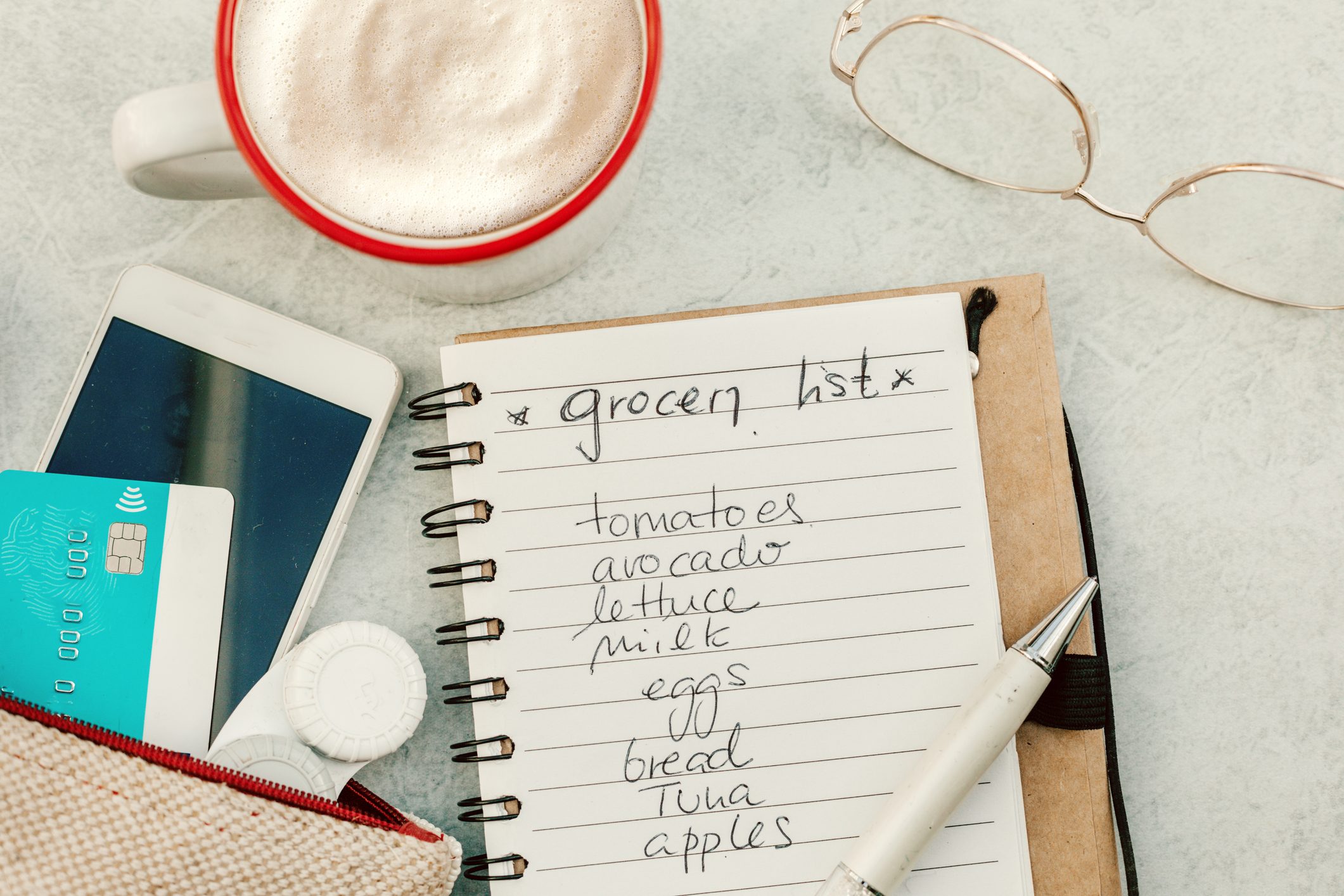 top view of grocery list, glasses, a purse with a credit card and phone and a mug of coffee