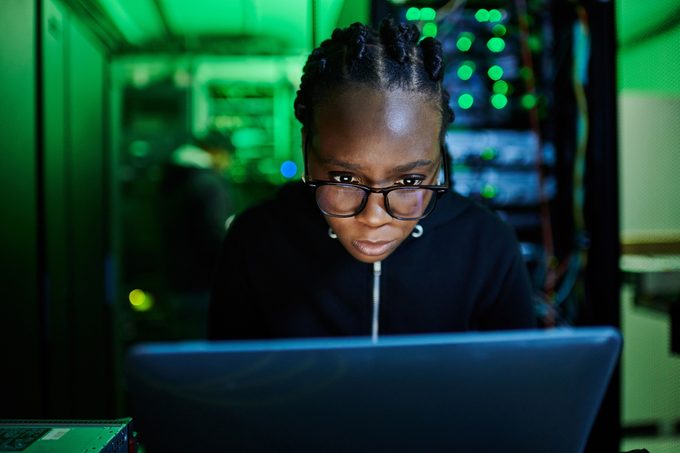 Person concentrates intently on a laptop in a dimly lit server room with green lighting and blinking equipment.