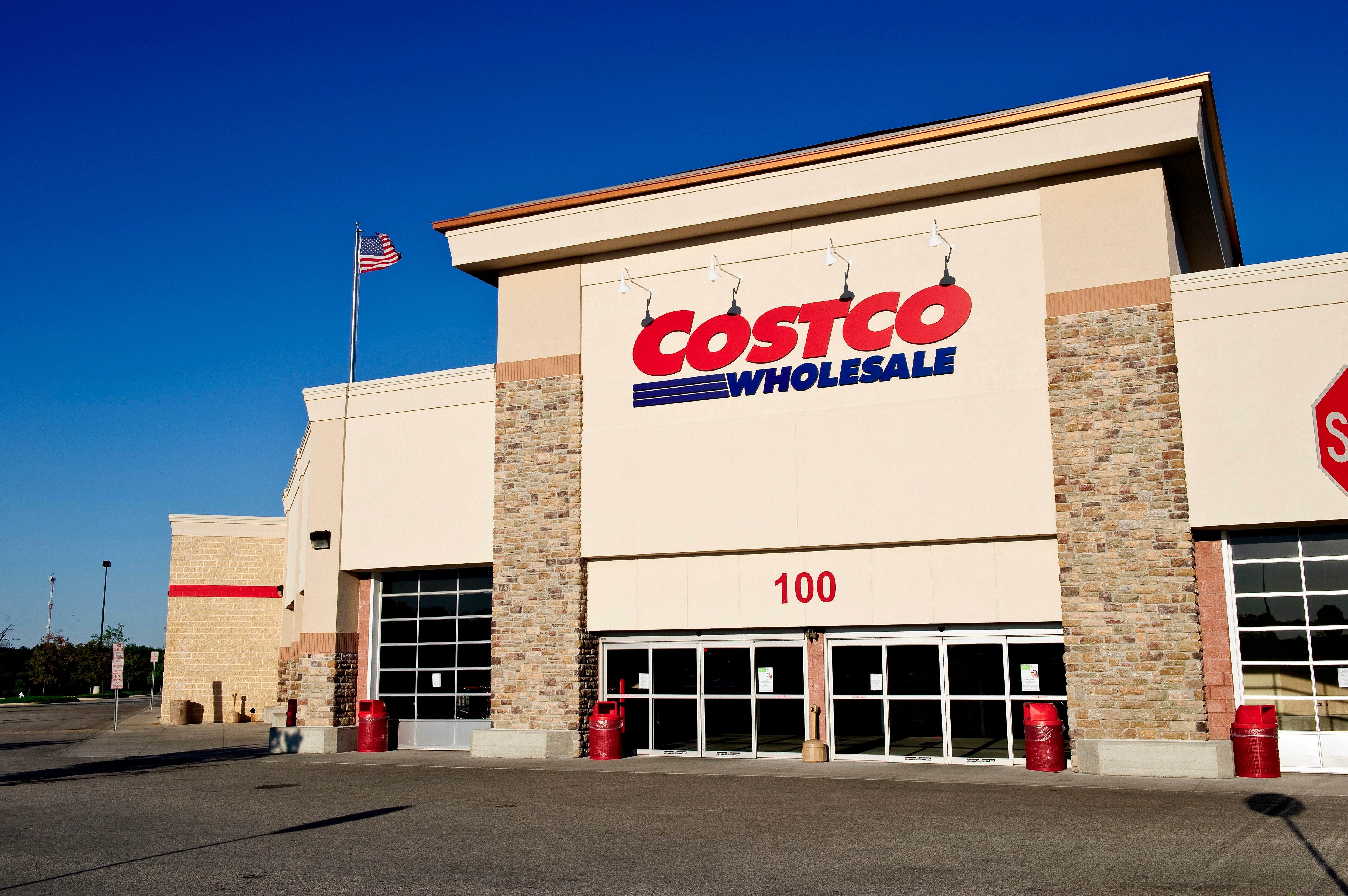 Building displays "COSTCO WHOLESALE" sign, featuring stone accents; American flag flies overhead against clear sky; empty parking lot and stop sign visible.