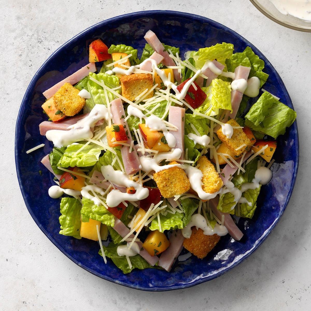 Salad with lettuce, ham strips, peaches, croutons, and shredded cheese, drizzled with dressing, sits on a blue plate against a light background.