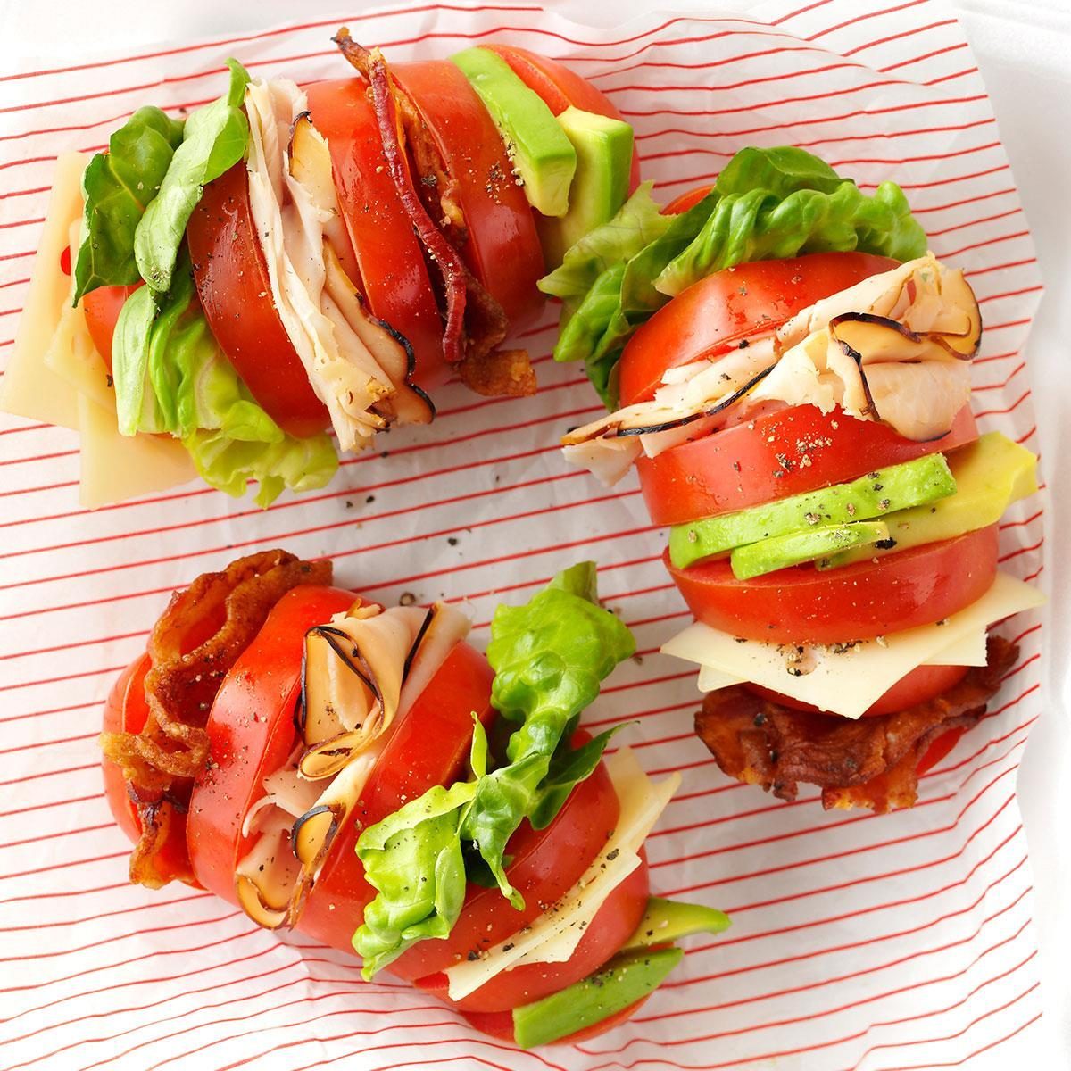 Tomato sandwiches layered with turkey, cheese, lettuce, avocado, and bacon sit on striped paper, showcasing a fresh, colorful arrangement.