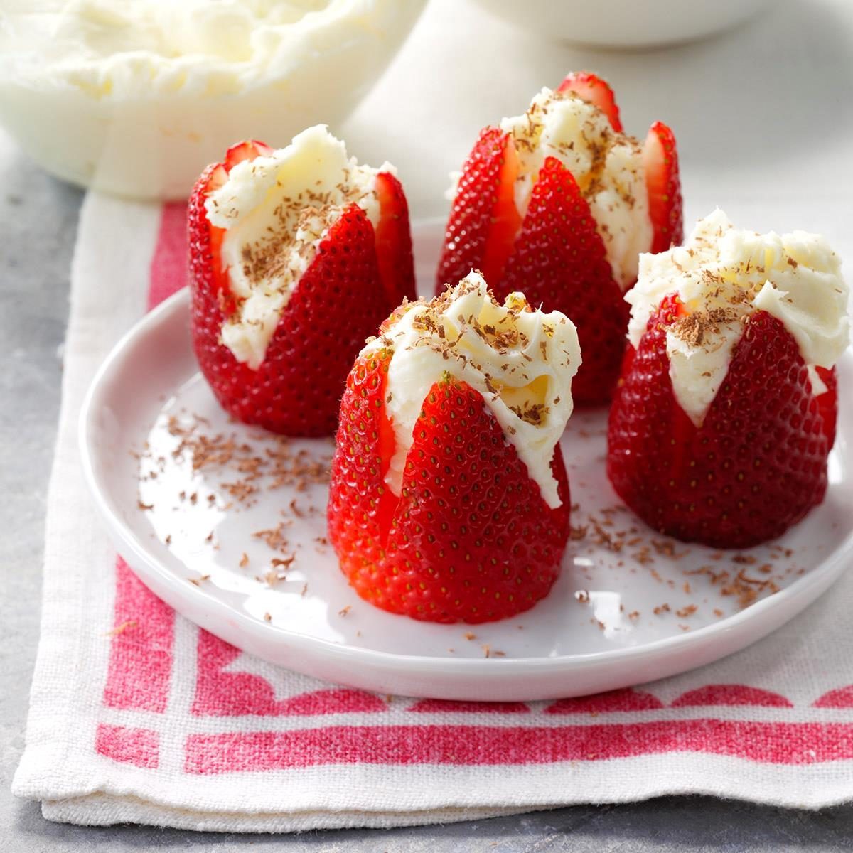 Strawberries filled with whipped cream sit on a white plate, sprinkled with chocolate shavings, over a red-and-white patterned cloth.