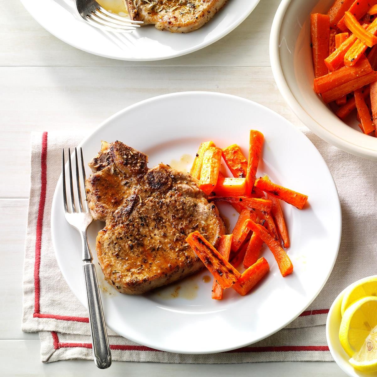 Grilled pork chop sits on a plate with roasted carrots, placed on a striped napkin, with a bowl of more carrots nearby.