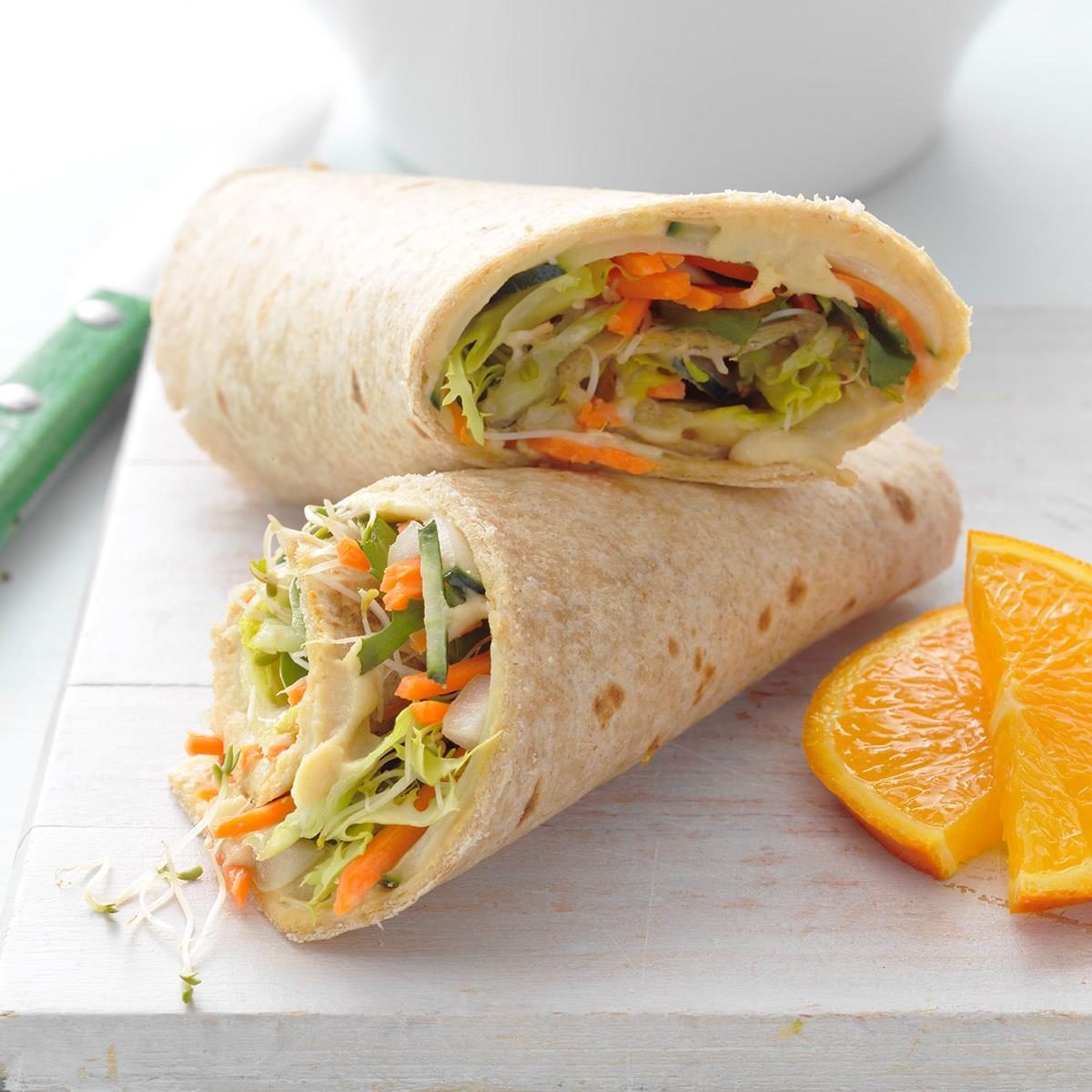 Wrap rests sliced on a white surface, showcasing vibrant vegetables inside; orange slices lie beside in a casual, fresh dining setting.