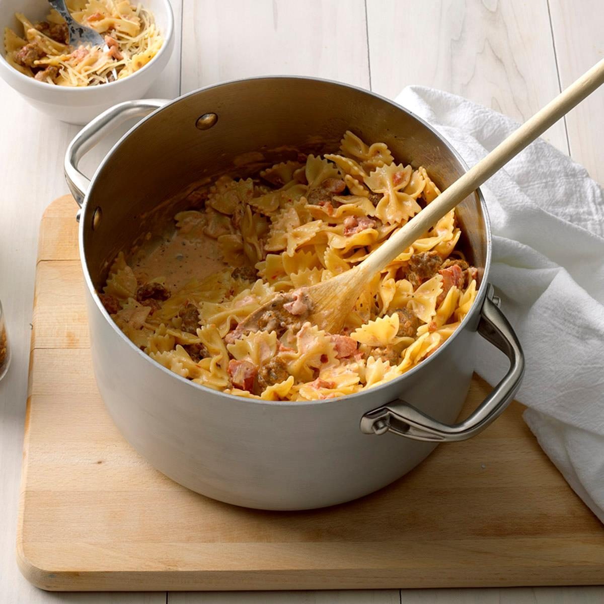 Pasta cooks in a large pot with a wooden spoon, resting on a wooden board, next to a bowl and white towel.