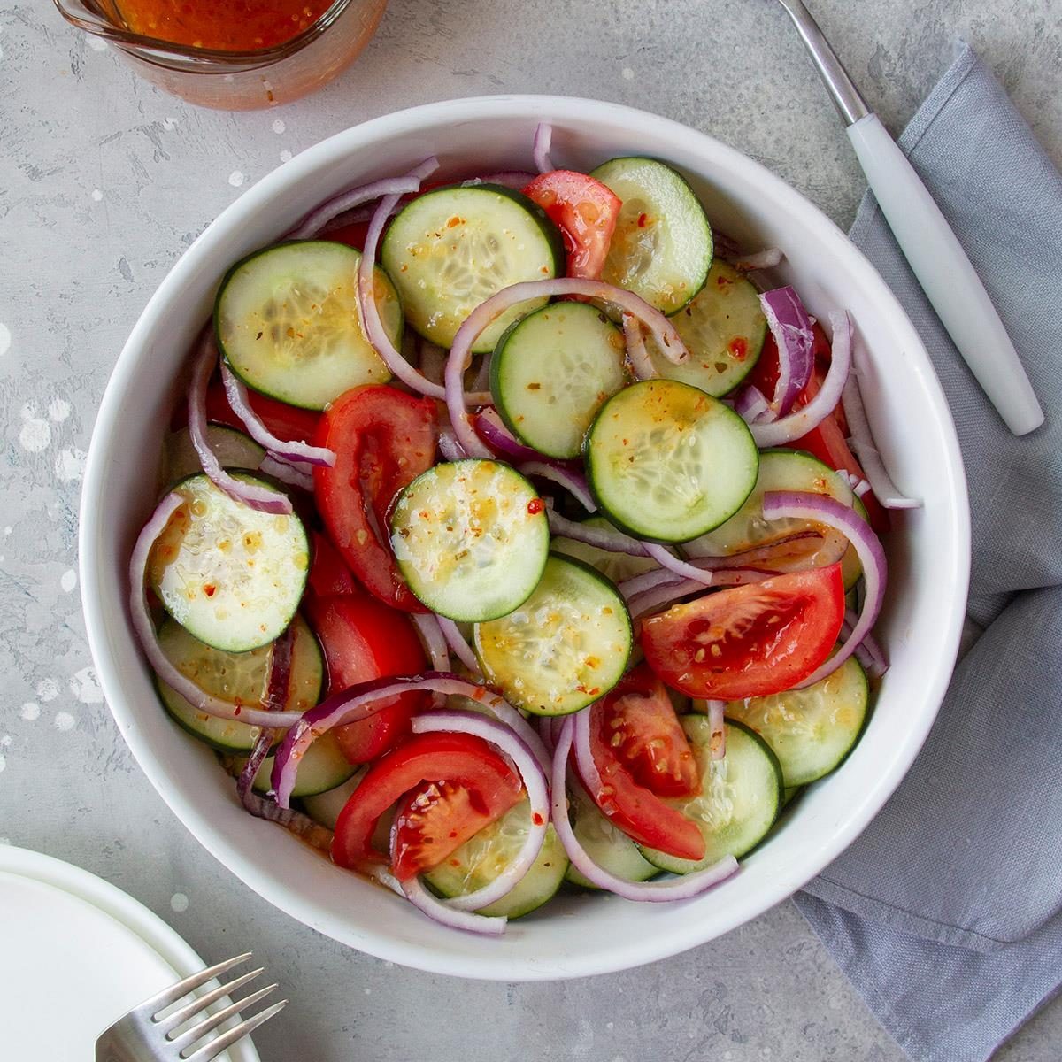 Salad features sliced cucumbers, tomatoes, and red onions in a white bowl, placed on a gray surface with a napkin, fork, and dressing nearby.