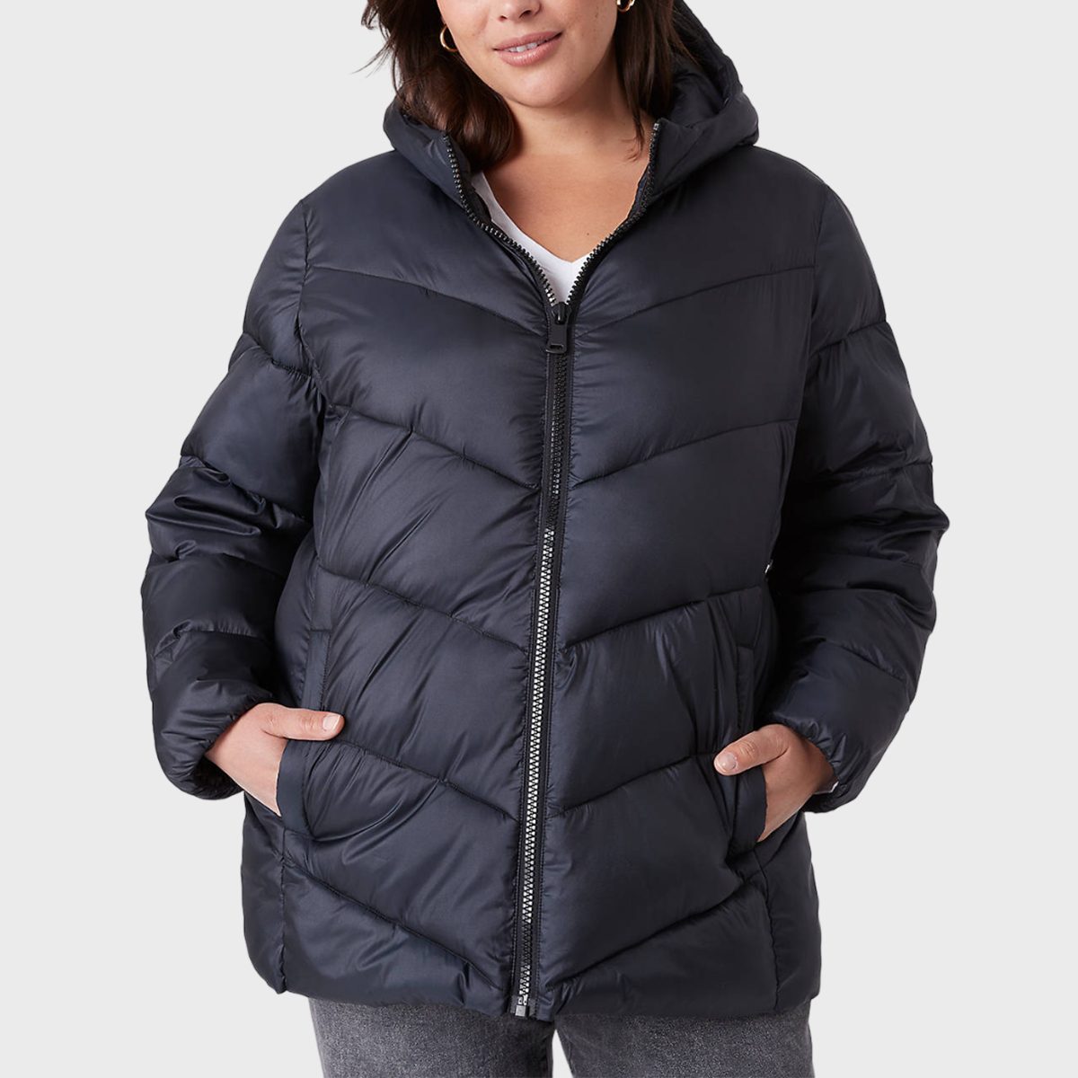 A person wears a dark puffer jacket, hands in pockets, against a plain background.