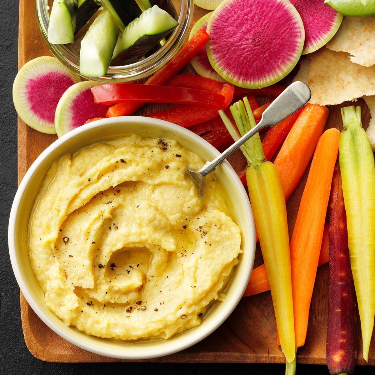 Bowl of hummus sits with a spoon, surrounded by sliced vegetables like carrots, cucumbers, radishes, and red bell peppers on a wooden board.