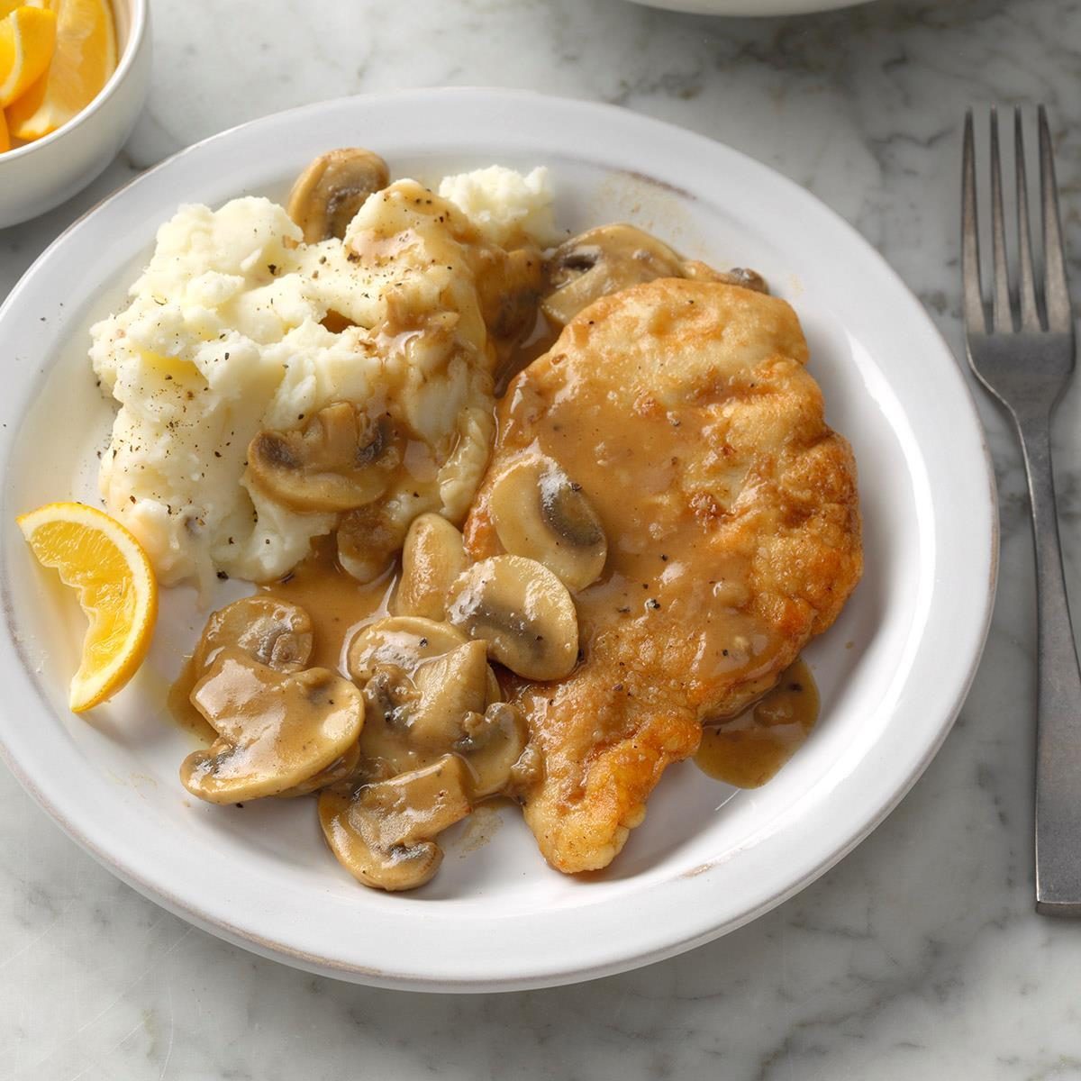 A plate holds breaded chicken with mushroom gravy next to mashed potatoes. A lemon wedge lies nearby on a marble surface.