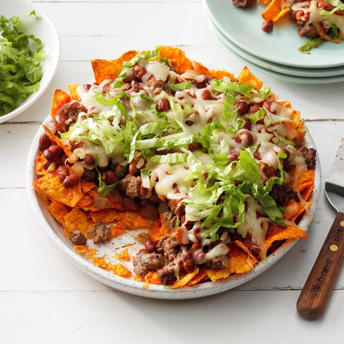Nachos topped with melted cheese, ground beef, beans, and shredded lettuce sit on a white plate beside a knife and other plates.