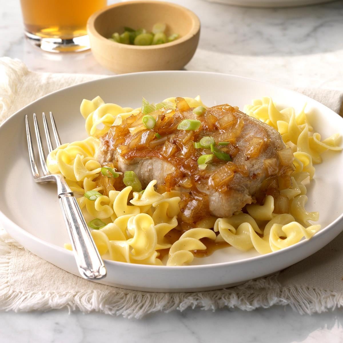 Pork chop topped with onions sits on egg noodles, garnished with green onions, beside a fork on a white plate with a light background.
