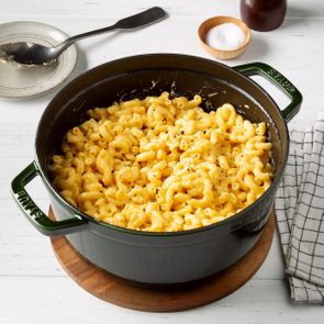 Macaroni sits in a pot on a wooden board, surrounded by salt bowl, spoon, and napkin on a white table.