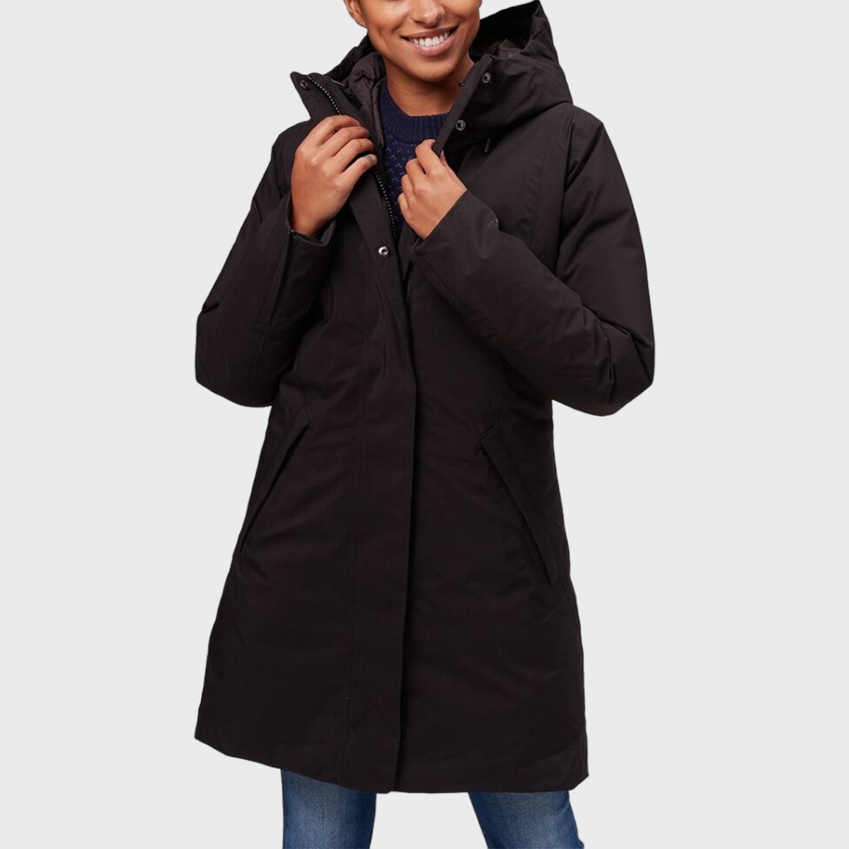 Person wearing a black hooded coat over a navy sweater, standing against a light gray background, with hands adjusting the front zipper.