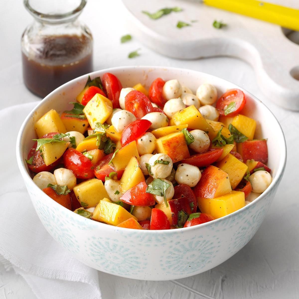 Salad combines cherry tomatoes, mozzarella balls, and mango chunks in a white bowl, with a small jar of dressing nearby on a white surface.