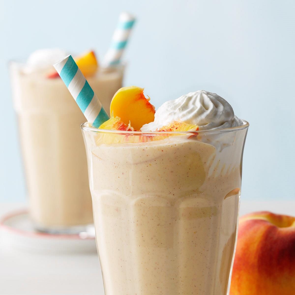 A peach milkshake with whipped cream and straw sits on a table, garnished with peach slices, against a light blue background.