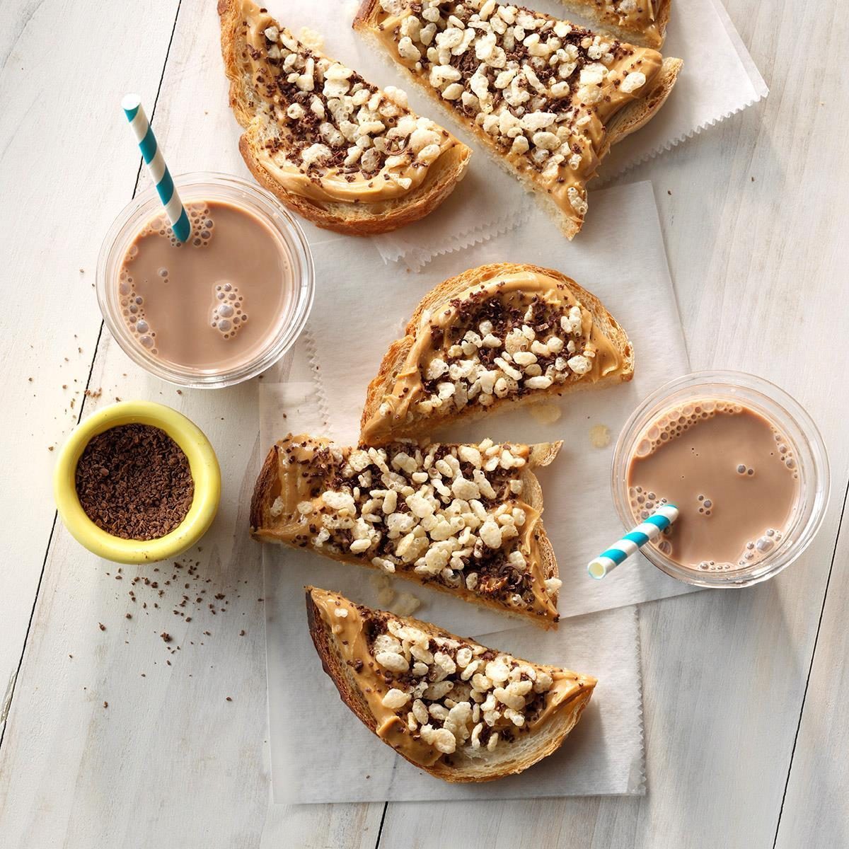 Toast slices topped with peanut butter and crispy cereal rest on parchment; served with chocolate milk in a cup with a striped straw.