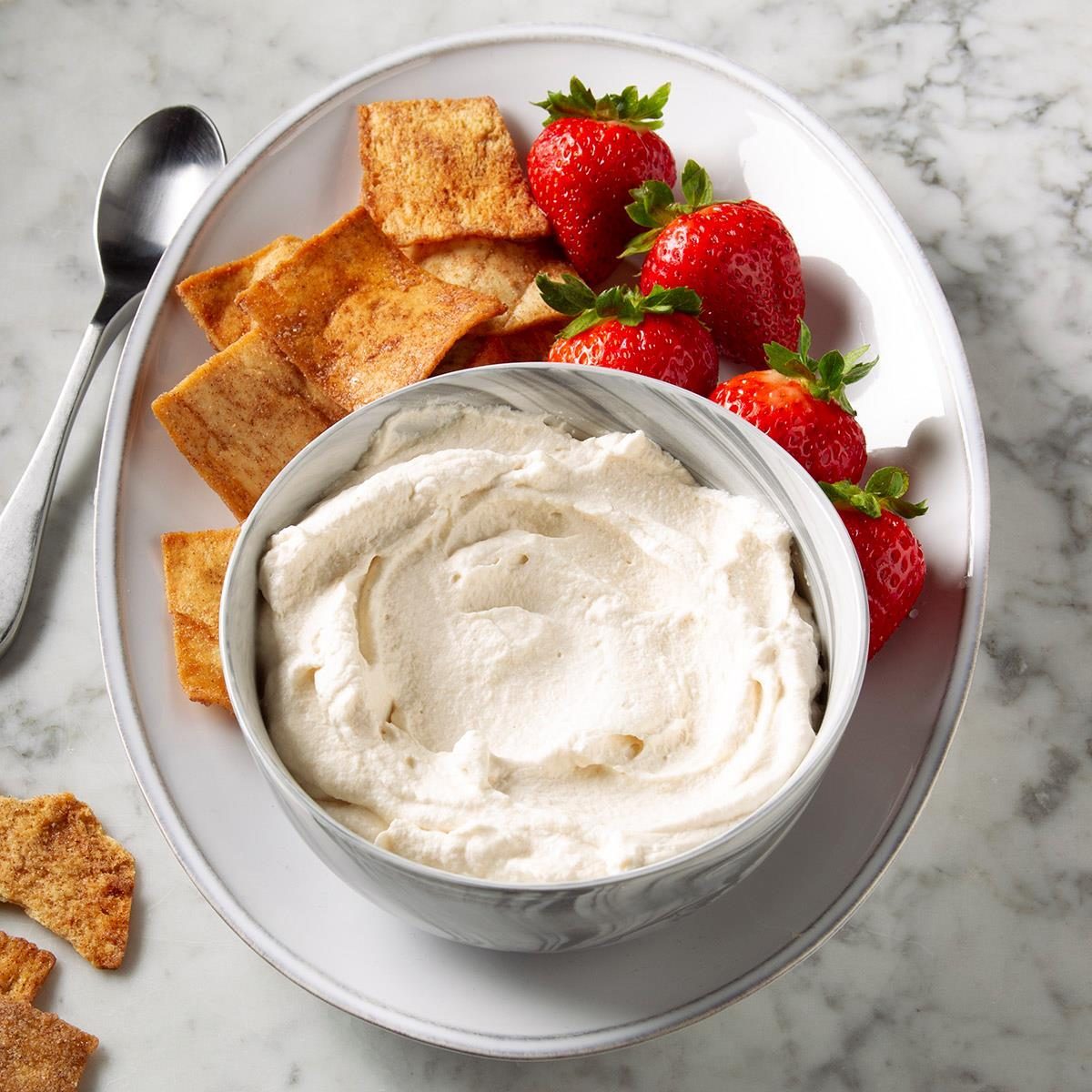 Bowl of creamy dip sits on a plate with strawberries and cinnamon-dusted crackers on a marble table, with a spoon nearby.