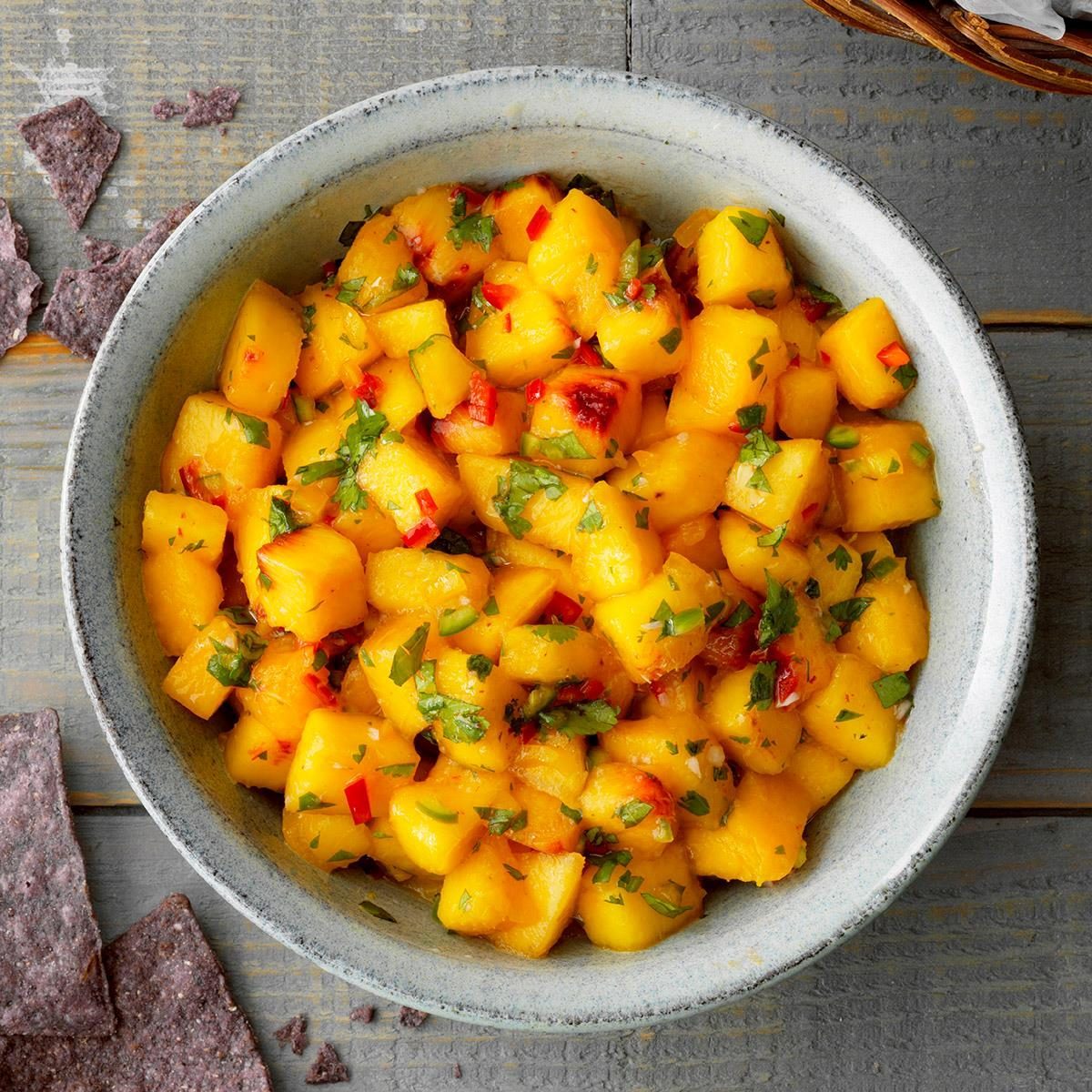 Mango salsa fills a bowl, garnished with red peppers and cilantro, beside scattered tortilla chips on a wooden surface.