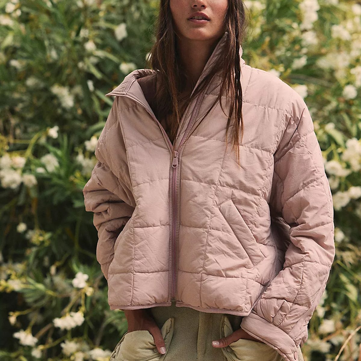 A person wears a light pink puffer jacket, standing in front of greenery and white flowers.