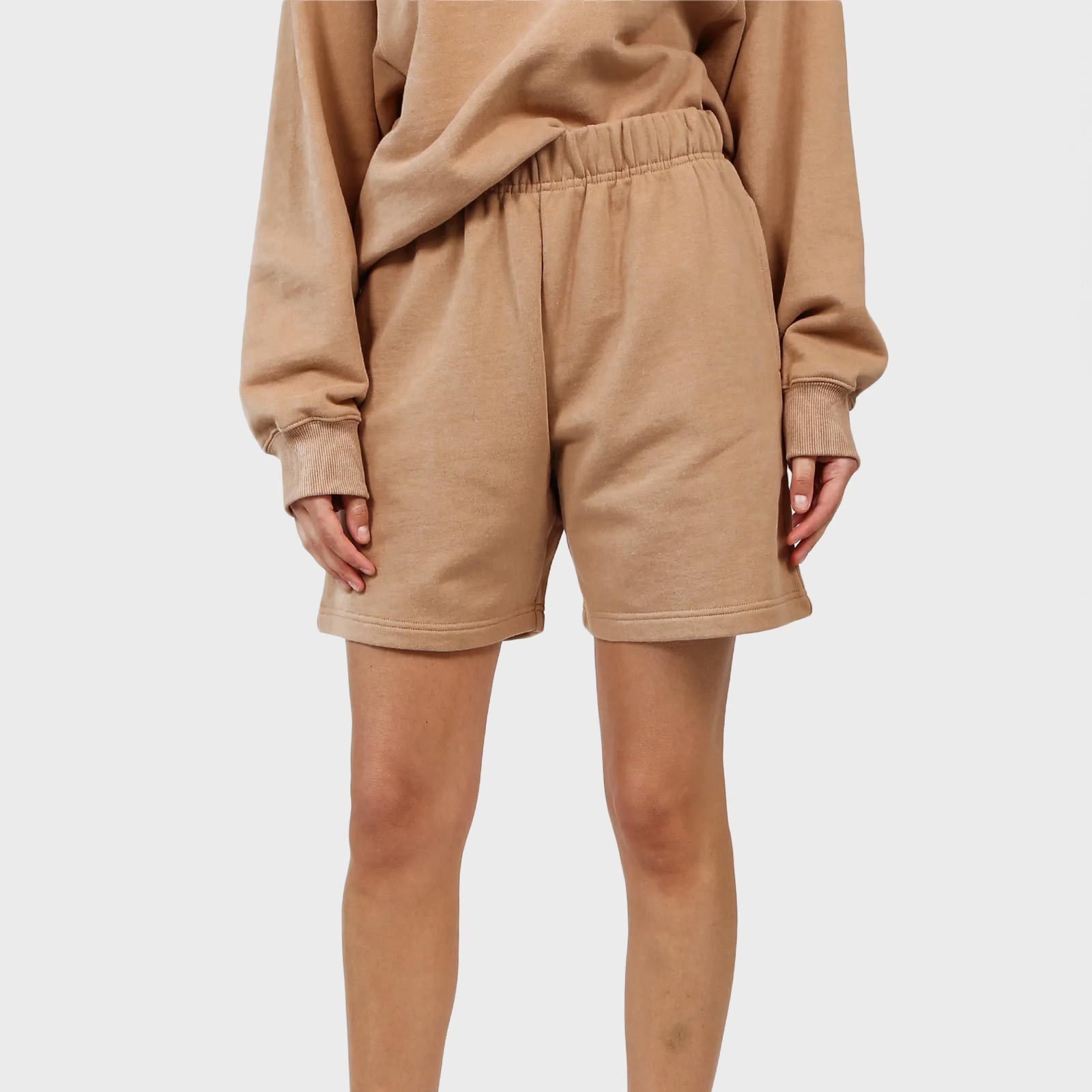 Beige shorts hang loosely on standing figure; matching long-sleeve top tucked in slightly. Plain white background.