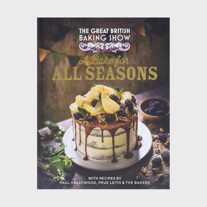 Cake topped with pears and blackberries, drizzled with caramel, sits on a wooden table. Accompanied by surrounding greenery and berries.