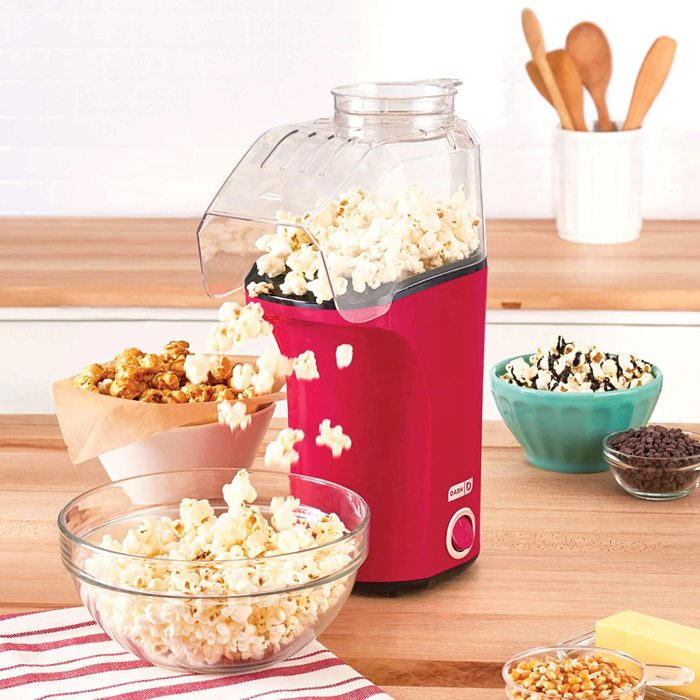 Popcorn maker ejecting fluffy popcorn into a glass bowl, surrounded by various snacks and utensils on a wooden kitchen counter.