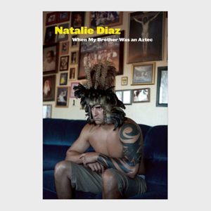Shirtless man wearing feathered helmet sits on couch, surrounded by framed pictures. Cover text: 