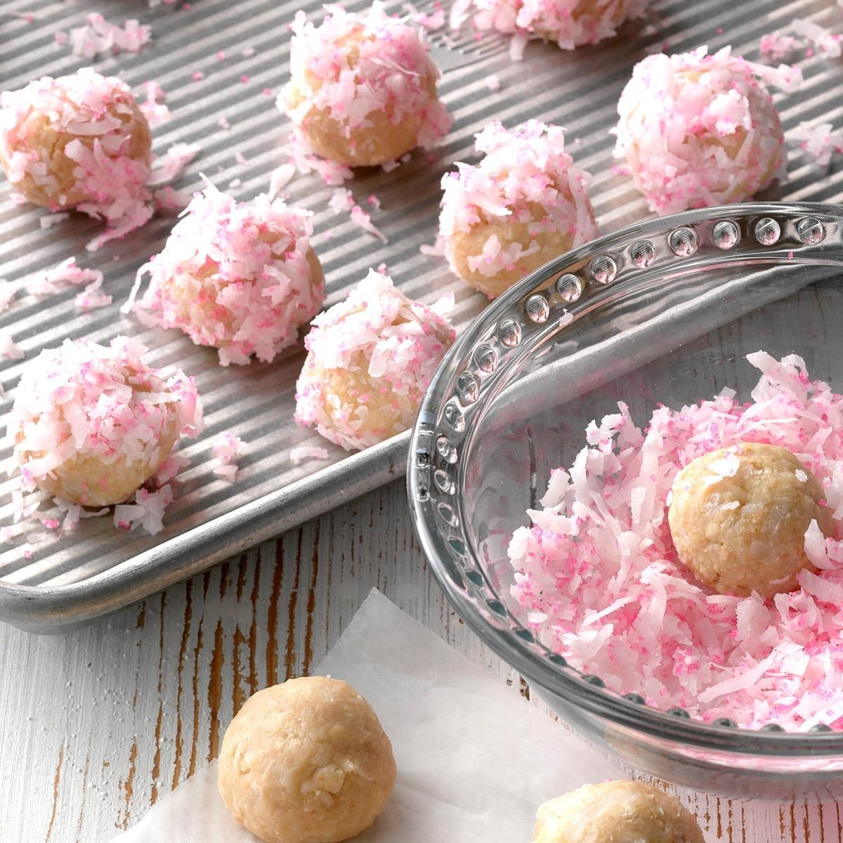 Coconut balls roll in pink coconut flakes, placed on a metal tray, with a mixing bowl and white wooden surface nearby.