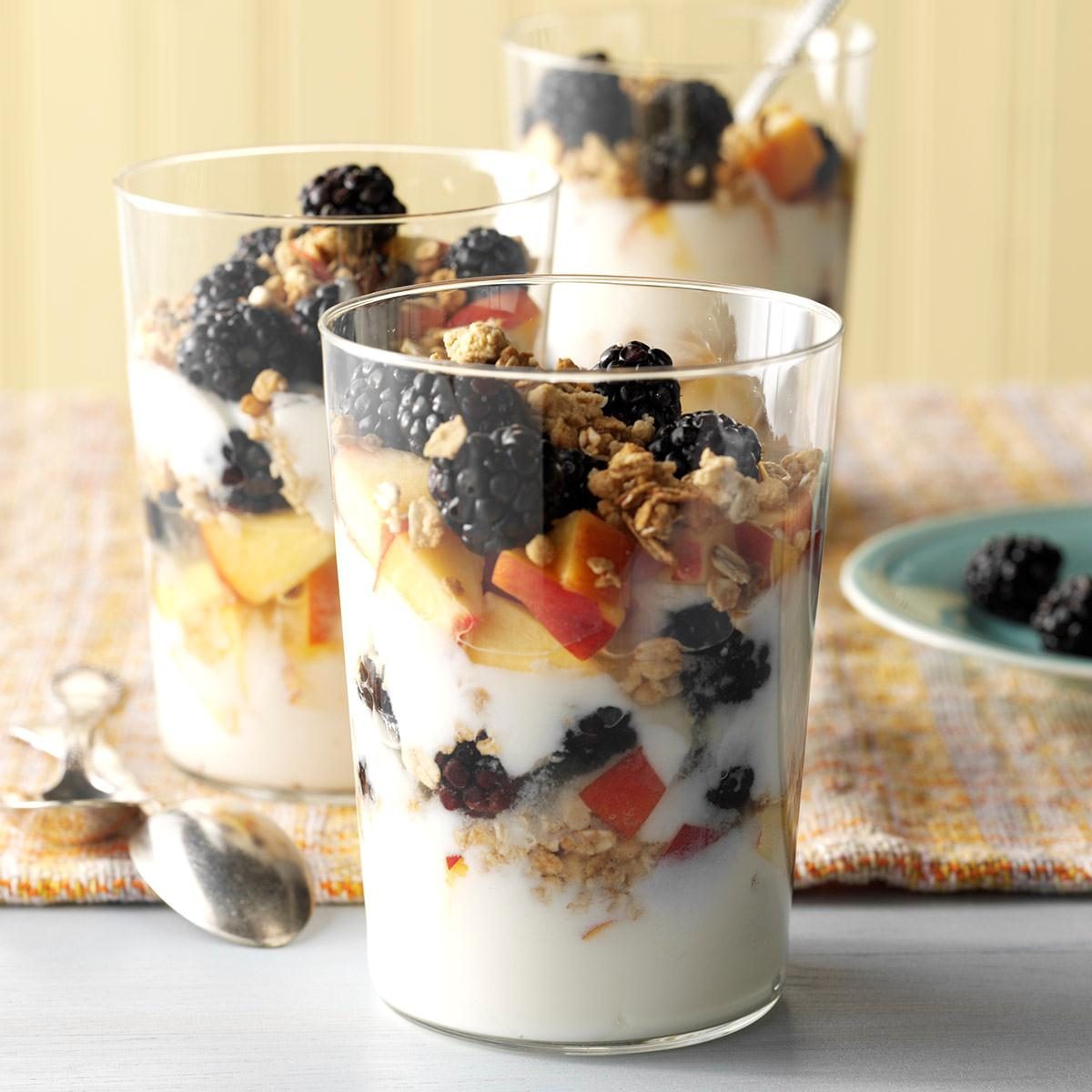 Glasses hold yogurt parfaits topped with blackberries and granola, resting on a table with a spoon and a plate of blackberries nearby.