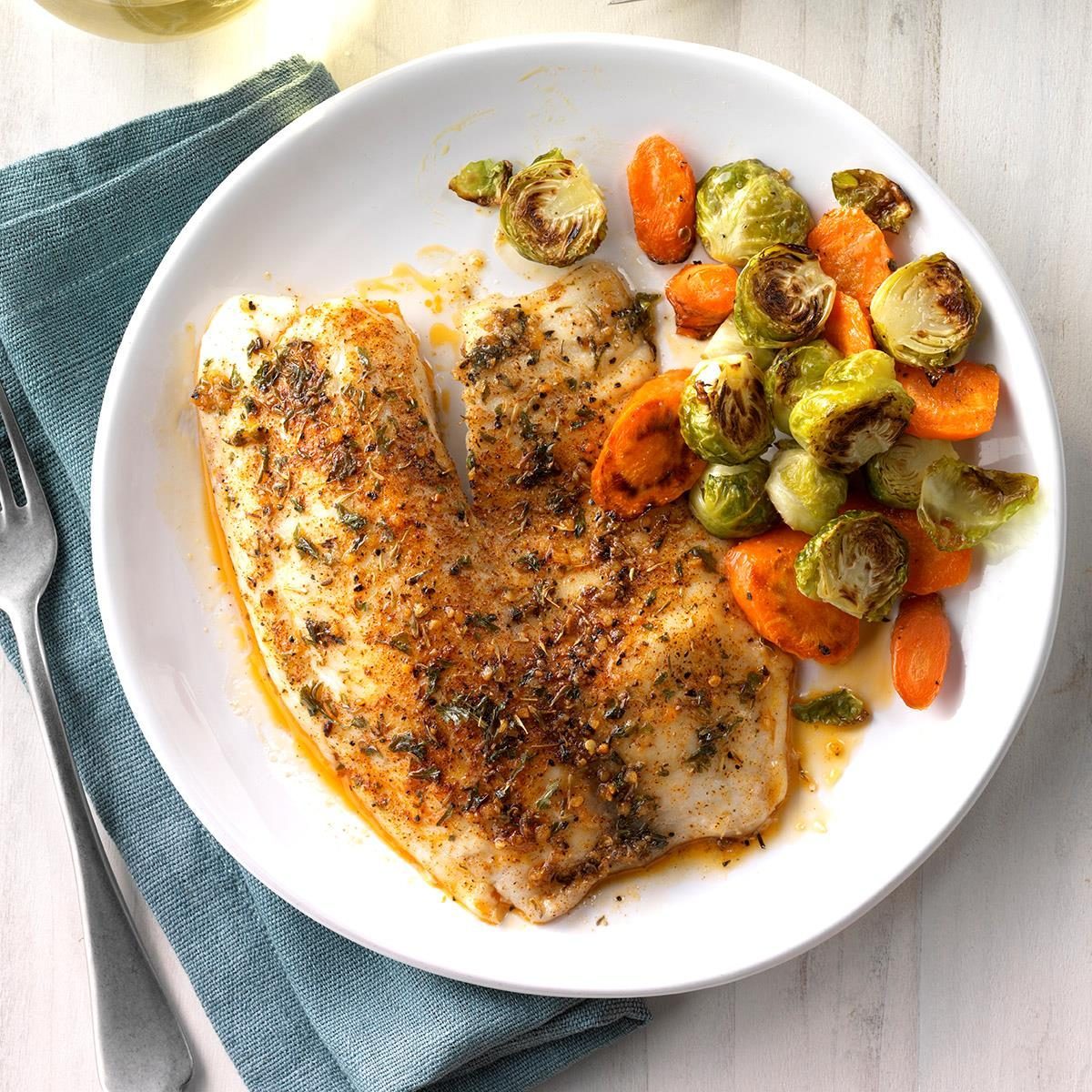 A seasoned fish fillet lies on a white plate beside roasted carrots and Brussels sprouts, atop a teal napkin with a fork nearby.