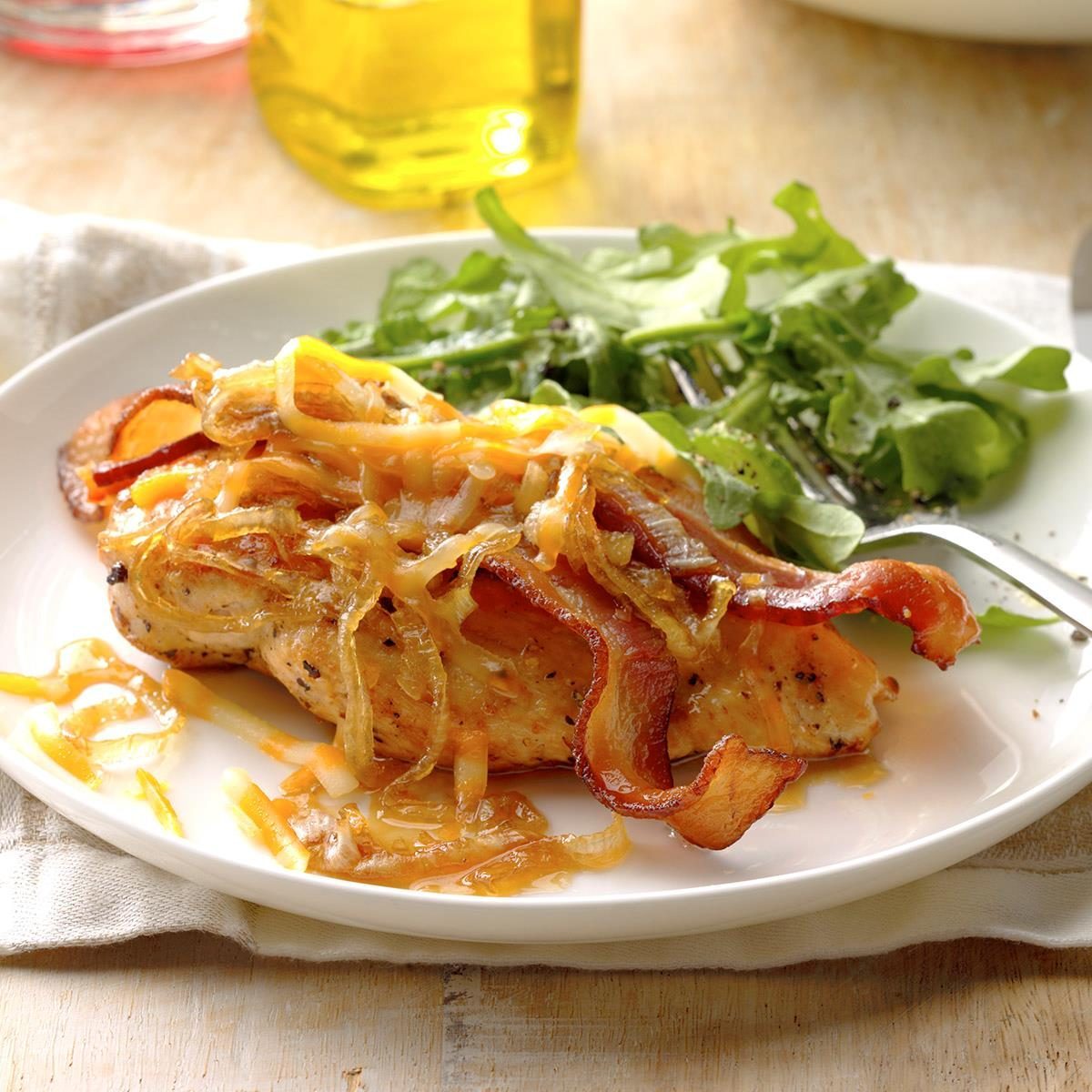 Grilled chicken topped with bacon and caramelized onions, accompanied by fresh greens, on a white plate in a rustic kitchen setting.