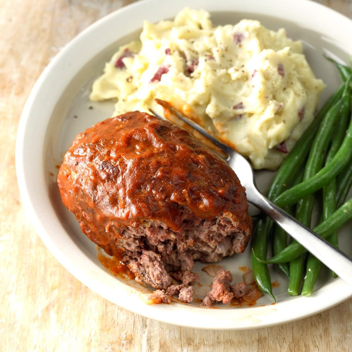 A piece of meatloaf sits next to mashed potatoes and green beans on a white plate, with a fork beside them.