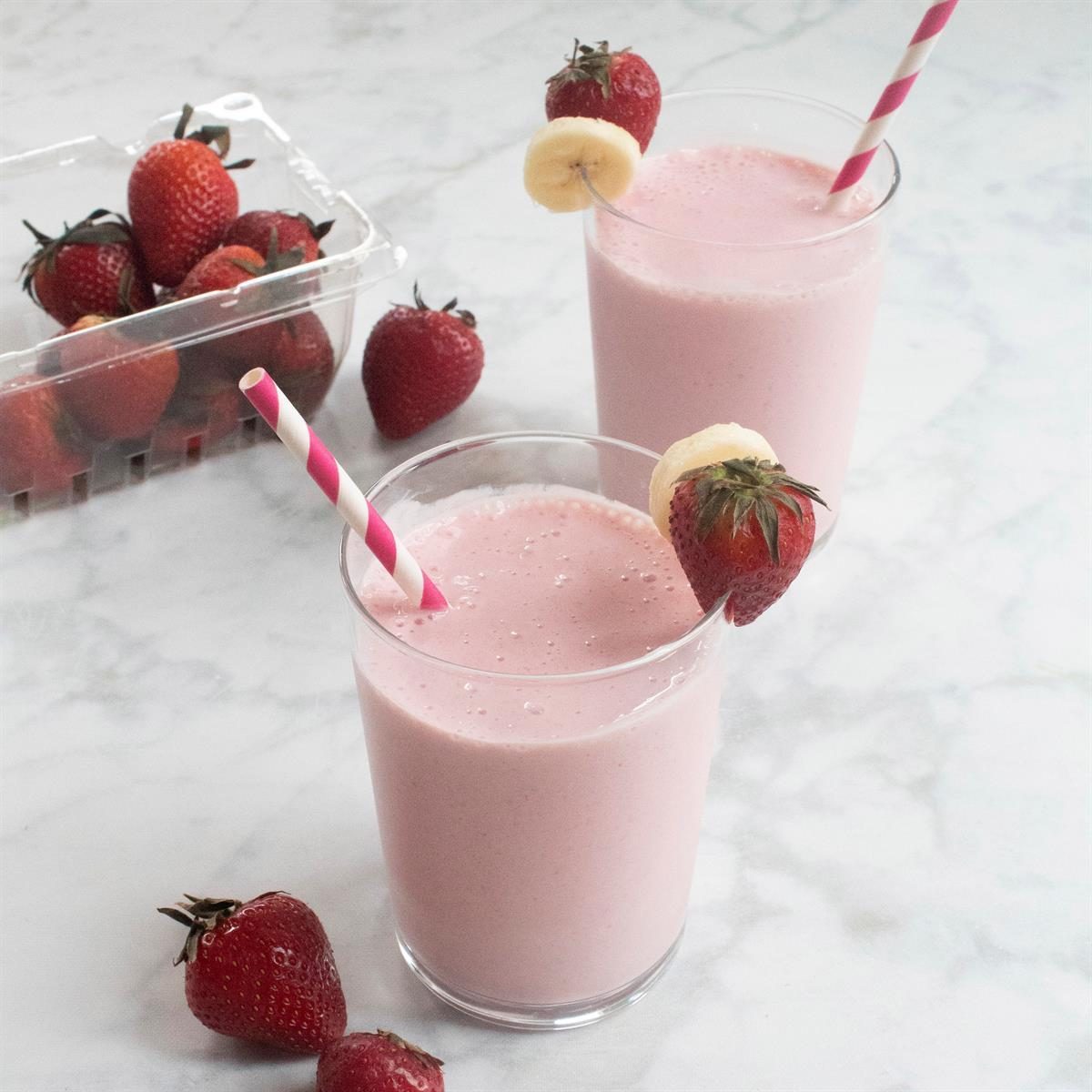 Smoothie glasses hold pink drinks with straws, garnished with strawberries and banana slices, beside a container of strawberries on a marble surface.