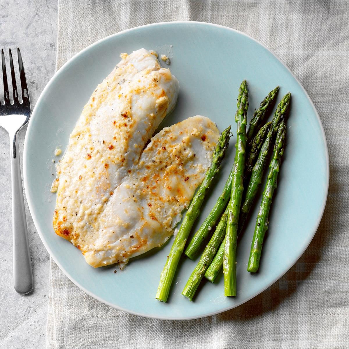 Fish fillets are served with asparagus on a blue plate, atop a plaid-patterned tablecloth, with a fork placed nearby.