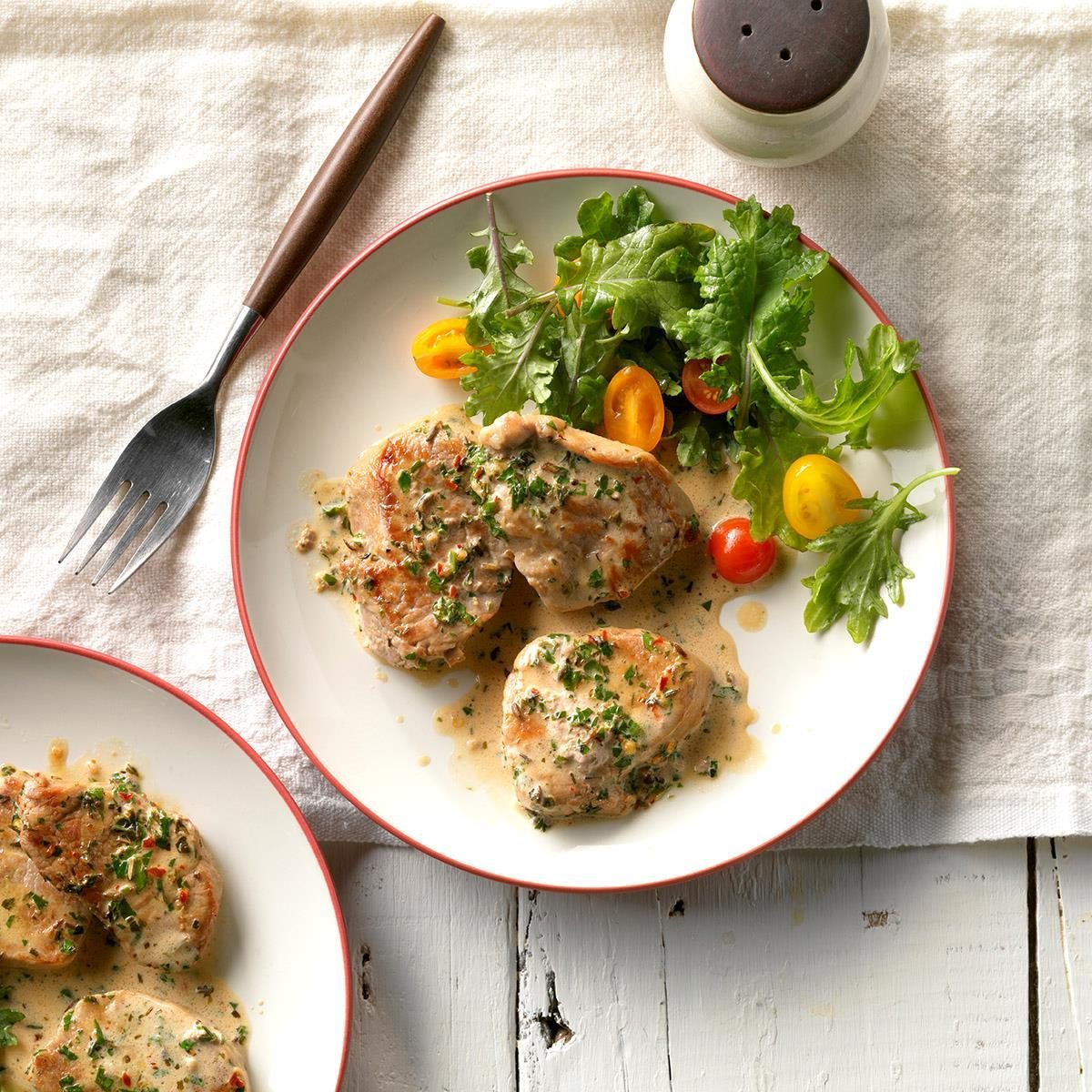 Plate holds creamy, herb-topped chicken paired with fresh salad on white linen, next to a fork and pepper shaker.