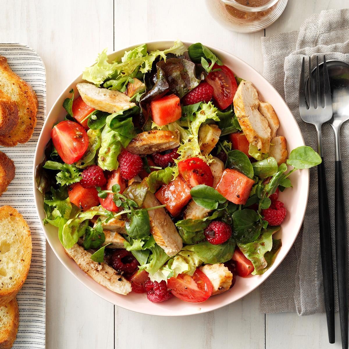 Salad sits in a bowl, combining grilled chicken, leafy greens, tomatoes, raspberries, and watermelon. Toasted bread slices rest nearby on a striped cloth.