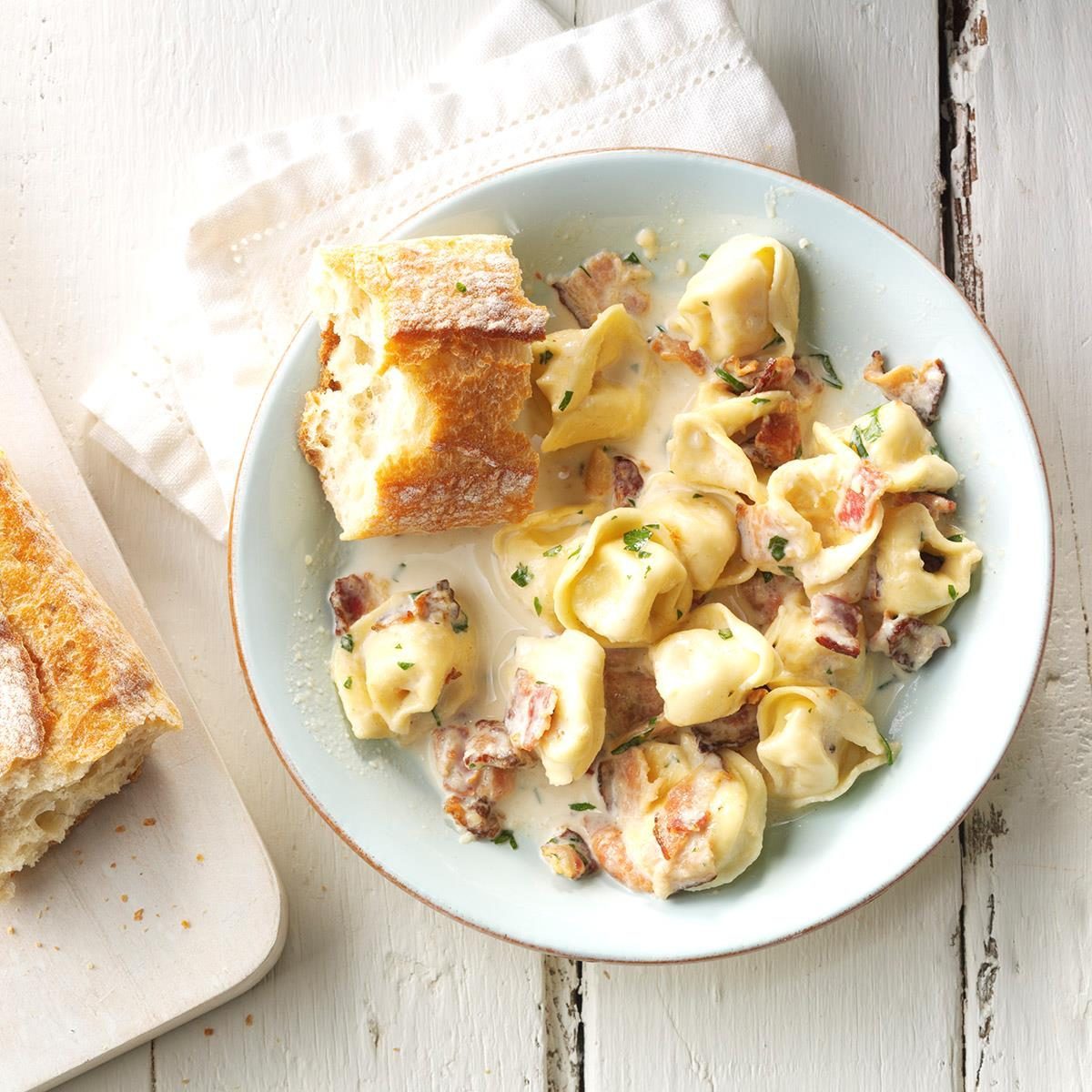 Tortellini with creamy sauce and bacon sit in a bowl, accompanied by crusty bread on a rustic white wooden table with a cloth napkin.