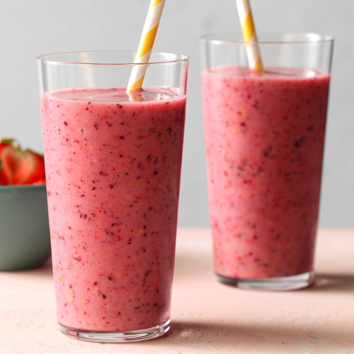 Two glasses contain pink fruit smoothie with straws, set on a light table, alongside a partially visible bowl of strawberries.