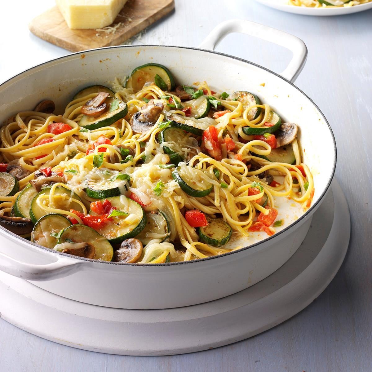 Pasta cooks in a white pan with zucchini, mushrooms, red bell peppers, and grated cheese on a kitchen countertop.